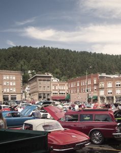 Historic vehicles parked haphazardly across a lot, many with their hoods open for a car show. Behind the parking lot is Historic Main Street, and pine and spruce trees on the mountain behind that.