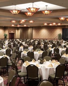 A convention center ballroom setup with round tables with eight chairs at each table. Tables are set with white table clothes and tan napkins.