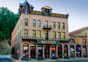 Tan brick three-story building with metal railings on upper-level balconies. Green sign with yellow lettering reads "Bullock Hotal." Located on Historic Main Street in Deadwood, SD.