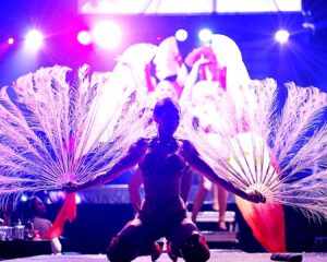 closeup of a woman on her knees on stage with red leather high heels boots on and holding large white peacock feather fans in each hand.