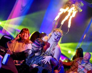aA blonde woman wearing a candelabra headpiece with three flames on top, seductively standing behind a sitting man dressed as a pirate.