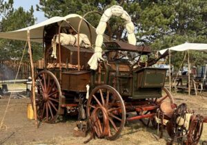 Deadwood's Chuckwagon Gathering and Cowboy Show with a picture of one of the historic and authentic wooden chuckwagons. the Wagon has a green body, red wheels and a canvas cover that is rolled up along the top.